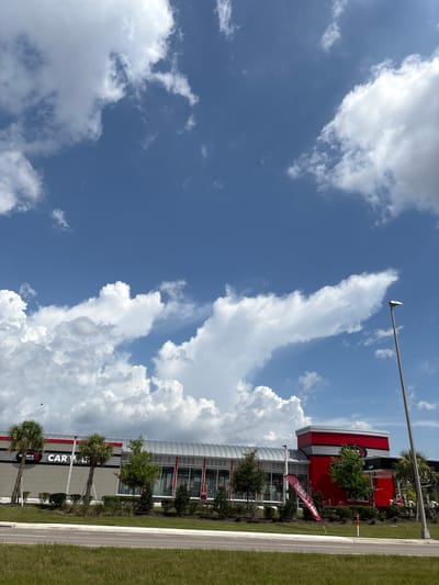 In Parrish, Florida, United States, a Tommy's Express Car Wash is captured under a bright, partly cloudy sky. The car wash features a modern architectural design with a long, silver, curved roofline and prominent red accents. The building has large glass windows and doors, suggesting an open and accessible space. In front of the building, lush green grass and well-maintained landscaping, including palm trees and various shrubs, line the road. A tall streetlight stands to the right of the building. The sky is a vibrant blue, dotted with large, fluffy white cumulus clouds that create dynamic shadows and highlights across the scene. One red flag with white text is planted in front of the building, and the words "CAR WASH" are visible on the building's facade.