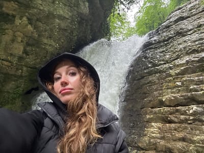The image shows a person wearing a black hooded jacket with blonde hair, facing the camera, with a waterfall in the background. The waterfall is flanked by rock walls and green vegetation. The photo was taken in Tsabal, Abkhazia. The image is upside down.