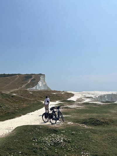 A person stands on a gravel path overlooking the Seven Sisters white cliffs in Seaford, United Kingdom. The individual, wearing a white t-shirt, beige trousers, and a dark cap, has a black cross-body bag. Two bicycles are parked on the path next to the person. The foreground is covered in green grass with scattered small white and purple flowers. The path leads towards the dramatic white cliffs, which are partially obscured by the terrain. Several other people can be seen walking along a path on the grassy hillside in the distance, leading up to the cliff edge. The sky is clear and blue.