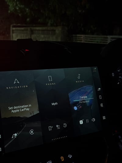The image shows a car's infotainment screen at night, displaying a menu with options for "Navigation," "Phone," and "Media." Navigation shows the text "Set destination in Apple CarPlay." Media displays "7 bitpib The Hardkiss," indicating a music selection. The car's dashboard is visible in the background, showing a speed of 0. The photo does not include a location in the city of Reis Magos, India.