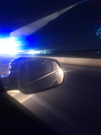 The image, taken at night, shows a view from inside a vehicle. A side-view mirror is prominently visible in the foreground, reflecting bright lights. The exterior of the vehicle, including a portion of the windshield and dashboard, is also visible. Bright blue and white lights are illuminating the scene from the left, suggesting the presence of emergency vehicles or other bright light sources. The background depicts a dark, open area, possibly a road or highway, with distant lights suggesting a town or cityscape. The location of Mingchinar, Uzbekistan, is not discernible from the visual information provided in the image.