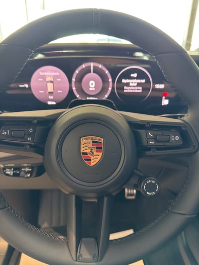 This is a close-up, eye-level interior shot of a modern car's steering wheel and dashboard, captured inside a dealership in Radinovo, Bulgaria. The steering wheel is black leather with visible stitching and has a central airbag cover featuring the Porsche crest. Various control buttons are visible on the steering wheel, including phone controls and media controls. The dashboard displays a digital instrument cluster with circular gauges showing speed in km/h, tire pressure in bar, and a clock displaying 15:55. A message on the display reads "Key/smartphone has been found. Please place in tray, see driver's manual." The overall lighting suggests an indoor environment, likely a showroom. There are no people or animals visible in the image.