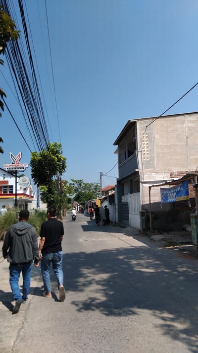 In the city of Bojongloa, Indonesia, a street scene unfolds under a clear blue sky. Two men, viewed from behind, walk away from the camera down a paved road. The man on the left wears a gray hooded jacket and blue jeans, while the man on the right wears a black t-shirt and blue jeans. The road is lined with buildings, including a multi-story structure with a gray facade and windows on the right, and smaller buildings with colorful accents on the left. A prominent sign for "CLIPPERCUT BARBERSHOP" is visible on the left. Trees, some with dense foliage, are scattered along the sides of the road. Further down the street, several other individuals can be seen, some walking and one on a motorcycle. Electrical wires crisscross overhead, casting shadows on the pavement. A blue banner with text and numbers, possibly advertising a sale or service, is hanging from a structure on the right side of the street. The overall atmosphere suggests a typical day in a residential or commercial street.