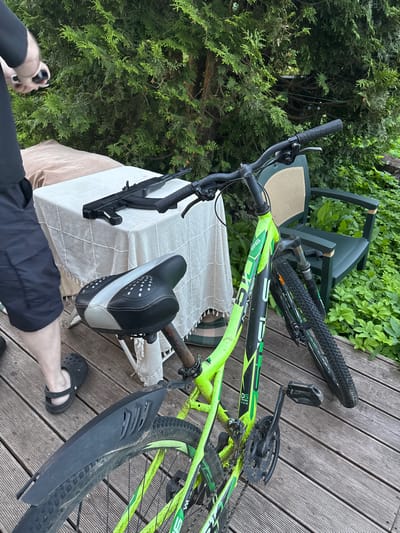 The image shows a bicycle parked on a wooden deck in an outdoor setting, likely a yard or garden. Behind the bicycle is a table covered with a light-colored cloth, upon which rests a black object resembling a rifle. To the right of the table, a green chair is visible. The background is dominated by lush green foliage, likely from trees and bushes. A person's legs, wearing black shorts and black footwear, are visible on the left side of the frame. The scene is outdoors, suggesting a residential area. The location, Воин Снт, Russia, is a horticultural non-profit partnership, commonly found in suburban or rural areas, which aligns with the visual cues of a garden and deck.