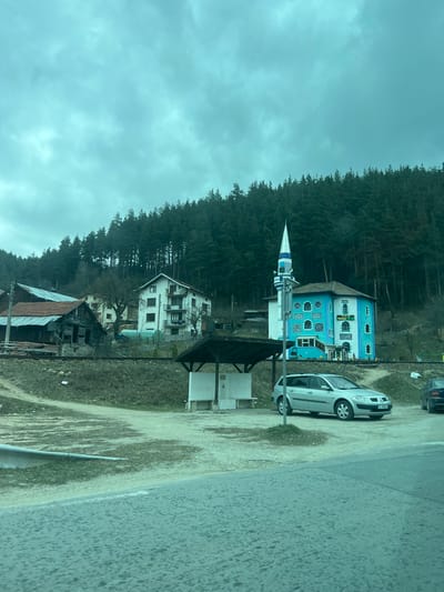 The scene, located in Cherna Mesta, Bulgaria, is observed under a heavily overcast, grey-blue sky. A dense coniferous forest covers a steep hillside that forms the background. In the mid-ground, from right to left, a two-story building painted bright turquoise with white ornamental script and white-framed windows is visible. Attached to its left facade is a slender minaret featuring a light blue and white striped conical spire, a blue cylindrical section, and a white base with a balustraded balcony. To the immediate left of this building, a multi-story white residential structure with balconies is situated higher on the slope. Further left and lower on the slope, a dilapidated wooden building with a rusted, reddish-brown corrugated metal roof is present, its structure appearing compromised. A single set of railway tracks runs horizontally across the mid-ground on a slight embankment. In the foreground, an unpaved, dirt and dry grass area transitions to a paved road segment in the lower right. A dark-roofed, open-sided shelter stands centrally in the unpaved area, housing two white, stone-like structures resembling public fountains or benches. A silver Renault Megane station wagon, with Bulgarian license plate 'C 1764 CY', is parked facing slightly right, adjacent to the shelter. A portion of a dark-colored vehicle is visible on the far right. No individuals are present, and no actions or interactions are occurring within the visible frame.