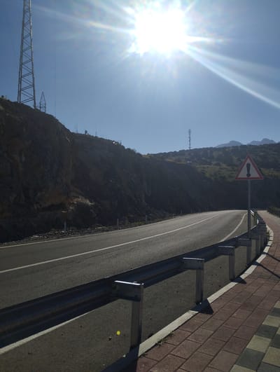 The image depicts a curving asphalt road with a guardrail on the right side, separating it from a brick-patterned sidewalk. The road is marked with white lines. To the left of the road is a steep, rocky embankment. In the background, there are hills and mountains under a bright, sunny sky with prominent sun flares. A tall, lattice-like tower is visible on the left side of the embankment, and another, smaller tower is visible on the crest of a hill in the distance. A triangular warning sign with an exclamation mark is also posted on the right side of the road, near the sidewalk. The scene appears to be an outdoor environment, possibly a mountain road. The location is identified as Myk, Uzbekistan. There are no people or animals visible in the image, and no discernible actions or interactions are taking place.
