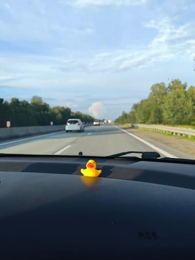 The image shows a view from inside a car driving on a highway. A small yellow rubber duck is placed on the dashboard in the foreground. The highway stretches into the distance, lined with trees on either side. The sky is blue with scattered clouds. Several cars are visible on the highway, and some traffic signs are present along the side of the road. The specific location in Barlak, Russia is not discernible from the image.