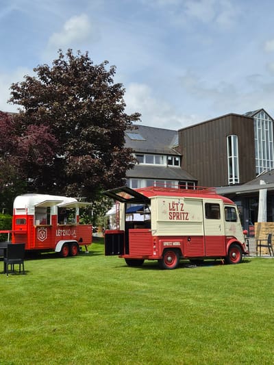 On a bright day in Lenningen, Luxembourg, the scene features two vintage red and white vehicles parked on a lush green lawn. The larger vehicle, branded "Lëtz Spritz," suggests a mobile bar setup. The smaller vehicle bears the name "Lëtz Kola". A building with modern architectural elements, including a glass-topped section, looms in the background. A dark-leaved tree stands beside the lower vehicle. There are no people visible in the immediate vicinity. The environment suggests a recreational or event space in the city of Lenningen.