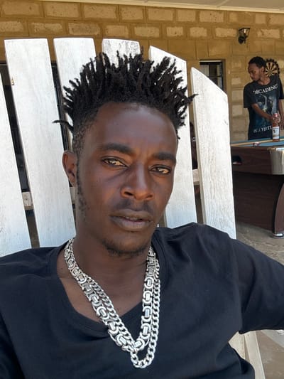 The image depicts an outdoor or semi-outdoor recreational area in Olkaria ward, Kenya, illuminated by bright daylight. In the foreground, a male individual with dark skin, short, upward-styled dark dreadlocks or twists, and minimal facial stubble is seated in a white, slatted wooden chair. He wears a black short-sleeved t-shirt and two distinct silver-colored chains around his neck, one appearing as a Byzantine-style link and the other a Cuban link-style. His light brown eyes are directed towards the viewer.
In the mid-ground, a second male individual with dark skin and short dark hair stands partially obscured by the foreground chair's slats. He is positioned in front of a light-colored brick wall and wears a dark t-shirt with white text, visibly displaying "DECADENCE" and "IS A VIRTU...". He holds a beverage bottle, consistent with a beer bottle, in his right hand. Directly in front of this individual is a blue-felted pool table. A dartboard is mounted on the brick wall to the right of the standing person, and a wall-mounted light fixture is visible above it. The ground surface is a light-colored paved area.