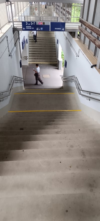 The image shows an outdoor staircase with concrete steps and yellow safety markings on some of the steps. There are metal railings on the sides of the stairs. In the background, there is a sign that reads "3 und 5-7", "Stadtmitte Waiblingen Süd", "Amts-/Bank" and icons for a toilet and a bus. A person is walking down the stairs carrying a bag. The environment appears to be a public space, possibly a train station or a pedestrian underpass, in Waiblingen, Germany.