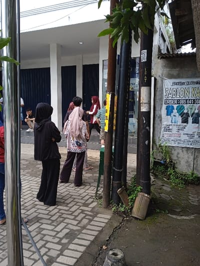 A street scene with people waiting outside a building with blue roll-down doors. A woman in a hijab and another carrying a child are prominent. Utility poles with signage and a poster for "FREEDAY CLOTHING - SABLON KA" advertising custom t-shirt printing services are also present. The street is paved with bricks in a herringbone pattern, and there is some greenery growing around the base of the poles.