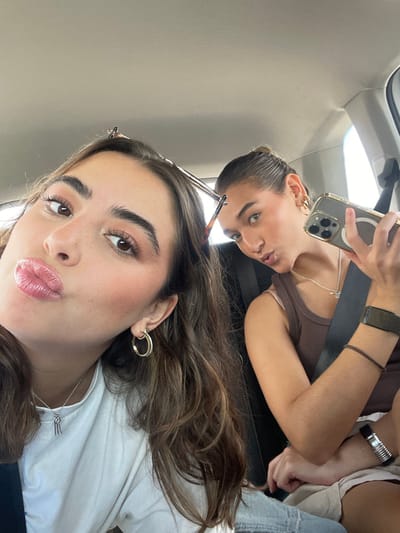Two young women are seated inside a vehicle in the city of Pedro Escobedo, Mexico. The woman positioned on the left, with long brown hair, wears a white top, gold-colored hoop earrings, and a silver necklace with a multi-bar pendant. She poses with pursed lips, looking towards the camera. A pair of sunglasses is perched on top of her head. The woman on the right, with dark hair pulled back, wears a brown tank top, gold-colored hoop earrings, a silver necklace, a dark wristband, and a metallic bracelet. She holds a gold-colored smartphone with a clear case and a magnetic accessory attached to its back in her left hand, angling it to capture a photograph. She also poses with pursed lips, looking towards the phone's lens. A dark seatbelt strap crosses her chest. The vehicle's light-colored headliner is visible above them.