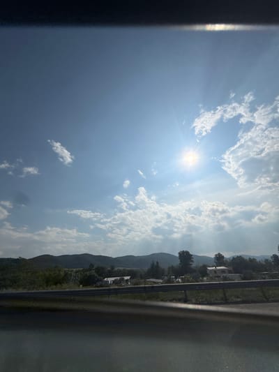 This image presents a wide, outdoor landscape view, consistent with a rural area in Mirovo, Bulgaria, captured from within a vehicle under bright daylight. The sky occupies the upper two-thirds of the frame, displaying a clear blue tone with scattered white cumulus clouds, which are denser on the right side. The sun is intensely radiant in the upper right quadrant, producing prominent lens flare and visible light rays. A dark, indistinct horizontal band is present along the top edge of the image.

Horizontally bisecting the lower third of the image is a metallic guardrail. Below this guardrail, the immediate foreground is dark and blurred, transitioning to a strip of green vegetation and dark ground. Beyond the guardrail, the midground reveals a landscape characterized by rolling hills covered in a mixture of dark green trees, lighter green foliage, and open fields. Several rural structures are discernible: a building with a light-colored or white roof is visible slightly to the left of the frame's center, and another, potentially older or disused building, is positioned further right. Additional smaller, indistinct structures and possible vehicles are clustered around these buildings. The distant background consists of a chain of darker, undulating hills or low mountains, partially softened by atmospheric haze. No people, animals, or specific actions are depicted.