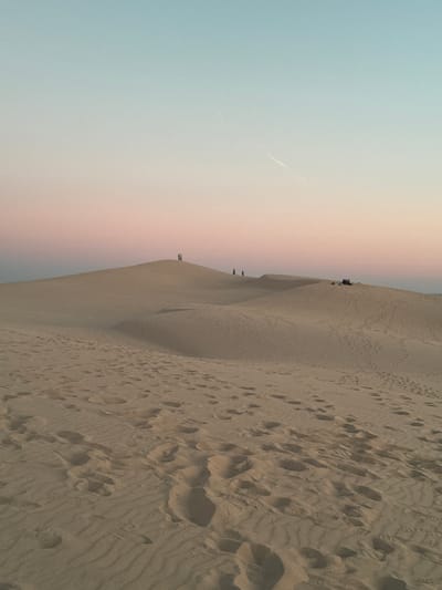 The image depicts a panoramic view of the Dune of Pilat, located in La Teste-de-Buch, France. The sand dunes stretch across the frame, with footprints visible on the sandy surface in the foreground. In the middle ground, several individuals are scattered on the crest of a dune, some standing and others appearing to be resting. The sky above transitions from a pale blue at the top to a soft pink and orange hue near the horizon, suggesting it is either sunrise or sunset. The overall scene conveys a sense of vastness and natural beauty.