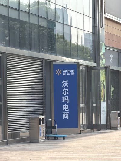 The image shows a modern building in Yuhuatai District, Nanjing, China. A large Walmart e-commerce sign is visible, mounted on the side of the building. The sign is blue with the Walmart logo and Chinese characters. The building has glass panels reflecting the surroundings. A metal waste disposal unit is attached to the building near the sign. The sidewalk is light-colored and runs along the building. No people are visible in the immediate scene.
