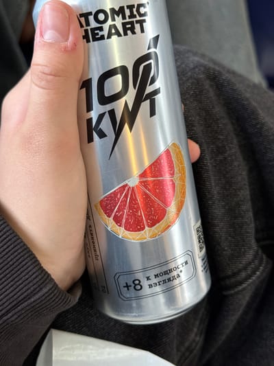 An individual's hand holds a silver aluminum can, branded with "ATOMIC HEART" and "100 KWT" in bold black lettering. A graphic of a grapefruit slice adorns the can, with segments highlighted in red and an orange rind. Below this, text in a white box reads "+8 к мощности взгляда*" which translates to "+8 to the power of gaze*". A QR code is visible on the right side of the can. The person is wearing a dark grey hooded sweatshirt. The background consists of dark fabric, possibly jeans or another dark textile. The scene appears to be indoors, with no discernible urban or environmental features of Aprelevka, Russia.