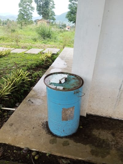 This image depicts an outdoor scene in Abbireddygudem, India. In the foreground, positioned on a damp, light grey concrete slab, stands a weathered, light blue metal drum. The drum's rusty lid, partially detached, collects standing water. The drum itself exhibits discolored patches and rust, with a faded rectangular mark on its side and a metal handle visible near the top. To the right of the drum, a vertical, off-white building wall or pillar extends upwards, showing dark stains and dampness at its base.
To the left of the concrete slab, a strip of cultivated ground features small green plants, some with variegated yellow-green leaves. Beyond this, a series of square, light-colored concrete pavers or stepping stones are arranged linearly within overgrown green vegetation. The ground adjacent to the concrete slab shows scattered dark, round objects, possibly fallen fruit or nuts.
The background reveals an expansive, unkempt green landscape dominated by numerous trees of varying sizes and a large patch of tall grasses. In the mid-distance, two small structures are visible: one light-colored building with a dark roof, and another with an orange or red roof further beyond. Distant hills or mountains rise under an overcast sky, completing the horizon. No individuals are present in the scene. The overall environment appears damp, suggesting recent precipitation.