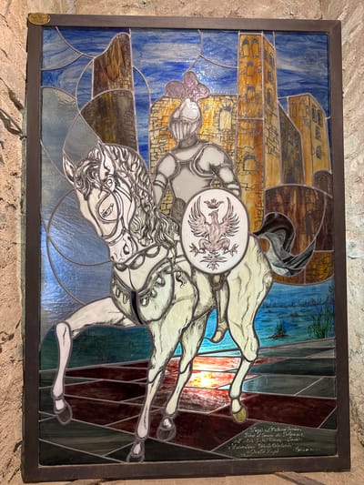 A stained-glass window depicts a knight on horseback in front of a castle. The knight wears armor and a helmet, and the horse is white with black accents. The knight holds a shield with a crest of an eagle. The castle is depicted with tall towers and arched windows. The background shows a blue sky with clouds and a body of water with reeds. The bottom of the window contains an inscription in Italian and a date, "Cortina 2003." The stained-glass window is located in Dolceacqua, Italy, at the Museum of the Castle of Dolceacqua.