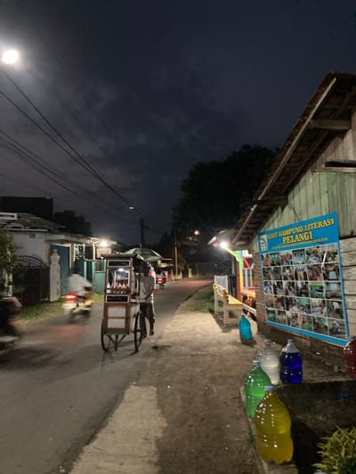 The image depicts a nighttime scene on a street in Proyonangg, Indonesia. A person is riding a pedicab, a three-wheeled passenger vehicle, which is illuminated by a nearby streetlight. The pedicab appears to be a food cart, with items displayed inside. Several large plastic bottles filled with colored liquids—blue, green, and yellow—are placed along the side of the road. In the background, there are buildings with a sign that reads "GKP KAMPUNG LITERASI PELANGI," indicating a "Rainbow Literacy Village" group. The sky is dark, suggesting it is late evening or night, and the weather appears to be clear. The overall atmosphere is that of a quiet, local street scene at night, possibly a marketplace or a residential area.
