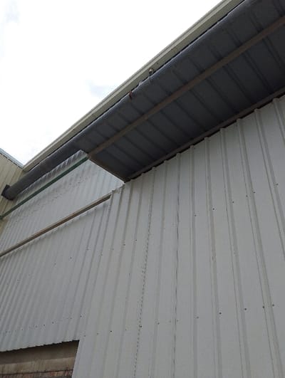 The image shows the exterior of a corrugated metal building in Pangasia, Bangladesh. The building is constructed of white, corrugated metal panels. A dark gray metal awning, supported by a metal frame, extends out from the building. The awning has a dark gray metal gutter along its edge. The gutter appears to be draining water. The building is in good condition. There are no people or objects in the image. The sky is overcast. The image is taken from a low angle, looking upward.