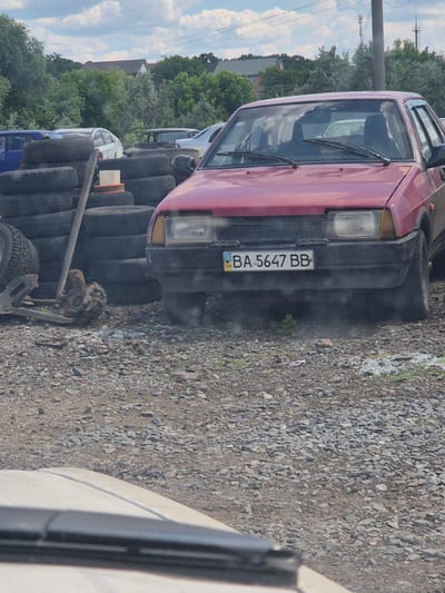 In Pidhaitsi, Ukraine, a red hatchback car with license plate BA 5647 BB is parked on a gravel lot, surrounded by stacks of used tires and car parts. The car's front bumper is dark and worn, with a yellowed headlight and a visible license plate holder. Behind the red car, other vehicles are partially visible, including a blue car and a white car. A few trees and a building with a reddish roof are seen in the background, under a cloudy sky. The scene appears to be an informal vehicle service area or junkyard.