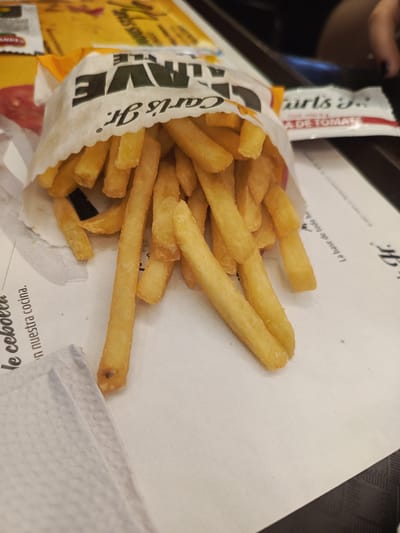 The image shows a serving of french fries from Carl's Jr. The fries are overflowing from a white paper bag with the Carl's Jr. logo prominently displayed. A packet of tomato sauce is visible in the background, also branded with Carl's Jr. The fries and bag rest on a white surface with Spanish text indicating the restaurant's place mats. Based on the logos and the place mats, this image probably comes from a Carl's Jr. restaurant. While there is no direct indication in the image, the Spanish text on the place mat and the name on the packet indicates this is likely from the Samborondón, Ecuador branch.