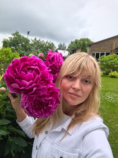 A blonde woman wearing a white jacket stands in a garden in Nikul'chino, Russia, holding a bouquet of vibrant pink peonies. She is positioned on the right side of the frame, with the flowers partially obscuring her face. Behind her, a wooden house with large windows and green trees are visible. The sky is overcast, and the grass is lush and green with scattered white flowers.