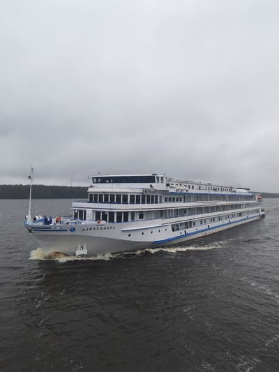 A large white passenger ship, named "ALEKSANDRA" in Cyrillic script, is moving across dark water under an overcast sky. The ship is several decks high, with numerous windows. The bow of the ship is creating a frothy wake. Trees line the distant shoreline. The location is in Topornya, Russia. There are three individuals visible on the bow of the ship.