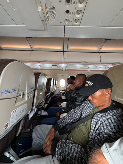 The image shows a row of passengers seated inside an aircraft, likely during a flight. The view is oriented along the length of the cabin, showing several rows of seats. The passenger closest to the camera is an older man wearing a black baseball cap with the word "STON" visible on it, along with a patterned jacket and a seatbelt. He appears to be sleeping or resting. Behind him are other passengers, all also seated. The seats are mostly a light tan color with seatback screens or panels. Overhead bins are visible along the sides of the cabin. An emergency instruction card is visible on the seat in the foreground. The image does not provide sufficient information to determine the location is Pajang, Indonesia.