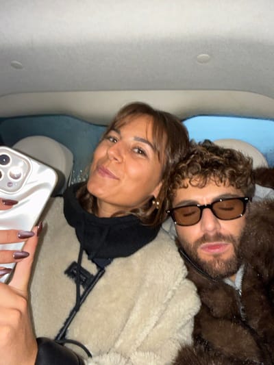 A woman holding a phone is posing with a man in what appears to be a car or airplane. The woman has brown hair and wears a black hoodie under a beige fluffy jacket. The man, wearing brown tinted glasses, has curly hair and a brown fur-lined jacket. They are close, potentially resting heads together. The phone being held by the woman has a distinctive white marble case. The background includes light-colored upholstery and a window with a glimpse of blue.
