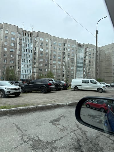 The image shows a street scene in Murmansk, Russia. A row of parked cars is visible along the right side of the street. The cars include a white van, several dark-colored SUVs, and a light-colored SUV. In the right side of the frame is a car side mirror, which shows a reflection of a red car. Behind the parked cars is a large, grey, Soviet-era apartment building with numerous windows. A street light pole is visible to the left of the building. The sky is overcast. The street surface appears cracked and worn.