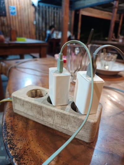 The image shows an electrical outlet on a wooden table in Geulumpangmeujim-Jim, Indonesia. Two white charging adapters are plugged into the outlet. Cables are connected to the adapters. In the background, there are tables and chairs with people sitting. Glasses and plates are visible on the tables. The environment appears to be an indoor space, possibly a cafe or restaurant.