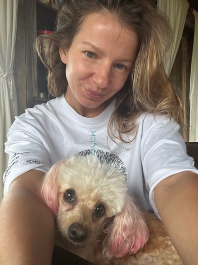 A woman with blonde, wavy hair and blue eyes, wearing a white t-shirt, smiles at the camera as she holds a light brown poodle with pink-dyed ears. The woman's right arm is visible holding the dog. The background consists of curtains and wooden structures, suggesting an indoor setting, possibly in Thôn 2, Vietnam. The lighting is natural, indicating it is daytime. The image appears to be a selfie.