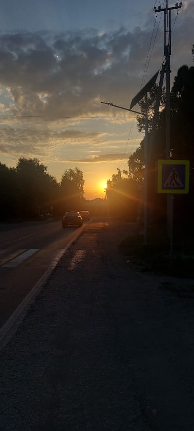 The image depicts a road scene at sunset in Berdsk, Russia. The sun is low on the horizon, casting a warm golden glow and creating lens flare. A pedestrian crossing sign is visible on the right side of the road. Trees line the left side of the road, with power lines and a utility pole overhead. Two cars are driving on the road, one in the foreground and another further ahead. The sky is a mix of blue and orange clouds. The overall mood is serene, with the warm light of the sunset creating a peaceful atmosphere.