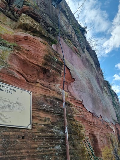 A low-angle shot depicts a weathered rock face with a metal pipe running horizontally across it. A blue wire parallels the pipe near the upper-left corner. The rock is predominantly reddish-brown with varying textures and some green moss or vegetation. A sign with text and diagrams is affixed to the lower-right of the rock face. The sign includes the words "Homburg" and "05-1714". The sky is visible in the upper portion of the image, showing a mix of blue and white clouds.