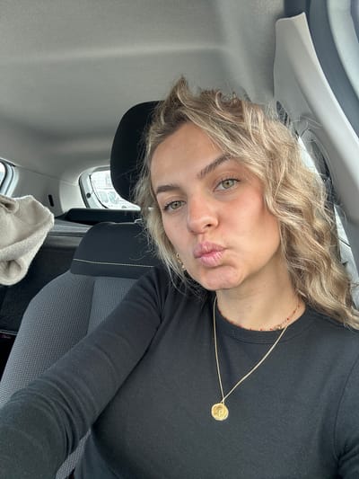 A light-skinned woman with blonde curly hair is taking a selfie. She has green eyes and is puckering her lips toward the camera. She is wearing a black long-sleeved shirt and a gold necklace. She is sitting in a car with light gray upholstery. The window to the left shows a blurred cityscape. The location is Valence, France.