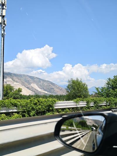 The image depicts a view from a vehicle traveling on a highway, likely near Nola, Italy. A tall cell tower stands prominently on the left side of the frame. In the background, a mountainous terrain with exposed rock faces is visible, partially covered by vegetation. Lush green bushes and trees line the side of the highway, creating a vibrant contrast with the rocky landscape. The sky is a clear blue with scattered white clouds. The reflection in the car's side mirror shows the highway stretching into the distance with other vehicles. The overall scene suggests a sunny day and travel through a rural or semi-rural area.