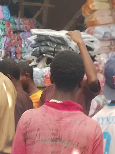 The scene is set indoors within a commercial establishment in Fagge D1, Nigeria, characterized by dense storage of merchandise. The background features shelving or stacked displays filled with numerous packaged items. On the left, rows of brightly colored objects resembling sandals or flip-flops, packaged individually in plastic, are stacked vertically in hues of green, red, purple, and blue. To the right, substantial piles of goods wrapped in black plastic are stacked, alongside further stacks of items wrapped in white or translucent plastic.
In the foreground, a person with short, dark, curly hair is seen from the back, wearing a short-sleeved pink collared shirt. The shirt's collar is a darker pink, and faint, dark markings are visible on the upper back. To the left of this individual, a person wearing a light brown shirt is partially visible from the back. Further left and slightly obscured, another person in a darker brown shirt is visible. Behind them, a glimpse of a person in a bright orange or yellow shirt can be seen. On the right side of the frame, another individual wears a dark blue baseball cap with an indistinct light-colored emblem and a white shirt with faded blue lettering on the back.
In the upper central portion of the image, a dark-skinned arm is raised, reaching towards the stacks of white-packaged goods in the background. The hand is positioned on or interacting with the top items of one such stack. No direct interactions between the visible individuals are discernable.