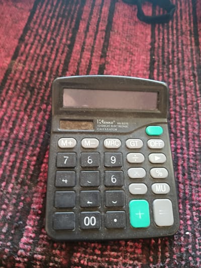 This media file features a black, 12-digit electronic calculator, specifically a KENKO KK-8378 model, positioned on a textured, red and black striped surface. The calculator has a digital display at the top and a keypad with numerical buttons (0-9), arithmetic operators (+, -, x, ÷), memory functions (M+, M-, MRC), a percentage key (%), a grand total key (GT), an off button, and an equals key (=). There is also a large green plus button. The scene is brightly lit, suggesting daytime, and there are no indications of specific weather conditions or a particular time of day beyond that. The setting appears to be indoors, possibly a desk or table, given the texture of the surface which resembles fabric, perhaps a tablecloth or a textured mat. No people are visible in the image. The visible text on the calculator includes the brand name "KENKO", model number "KK-8378", and the description "12-DIGIT ELECTRONIC CALCULATOR". The location context is Алат, Uzbekistan, though this is not directly inferable from the visual content of the image itself. The overall impression is one of an object placed in a static setting, with focus solely on the calculator and its immediate surroundings.