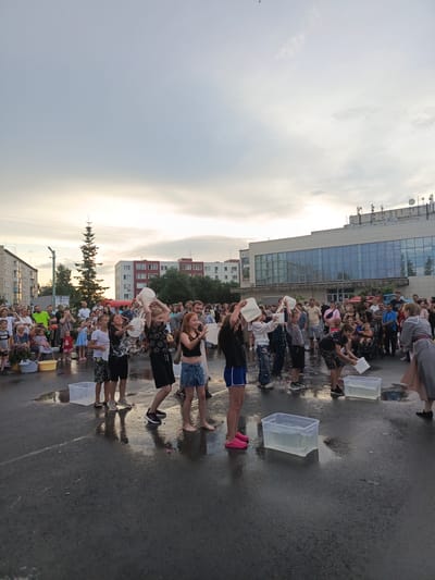 The media file shows an outdoor event in Криводановка, Russia, with many people gathered. In the foreground, several children are participating in what appears to be an ice bucket challenge or a similar water-drenching activity. They are pouring buckets of water over their heads, creating splashes. They are dressed casually in t-shirts, shorts, and skirts.
Surrounding the children is a crowd of onlookers of all ages, some holding phones to capture the moment. The event is taking place on a paved surface that is wet and reflective from the water being splashed. In the background, there are residential buildings and a modern-looking building with a large glass facade, possibly a community center or a public institution. A tall evergreen tree stands to the left of the frame.
The sky is overcast with hints of sunset, suggesting it is late afternoon or early evening. The overall atmosphere seems to be one of fun and community engagement, with participants and spectators appearing to enjoy themselves. There are several large translucent tubs filled with water scattered on the ground.
A sign with Cyrillic text, reading "РЕHДА" (RENDA), is visible on a building in the background, indicating a commercial or rental service. Another sign with Cyrillic text is present on the leftmost building. The weather appears to be warm enough for such an event, despite the cloudy sky.