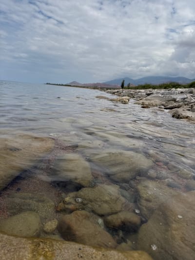 The image depicts a rocky shoreline with clear, shallow water lapping over smooth stones. In the foreground, submerged rocks are visible beneath the gently rippling surface of the water, with small bubbles appearing here and there. The shoreline is comprised of larger, greyish-brown stones. In the middle ground, a thin strip of dry land with sparse vegetation can be seen, leading to a distant mountain range under a cloudy sky. The scene appears to be a natural landscape, possibly a lake or a slow-moving river. The location is identified as a body of water in the city of Tong, Kyrgyzstan.