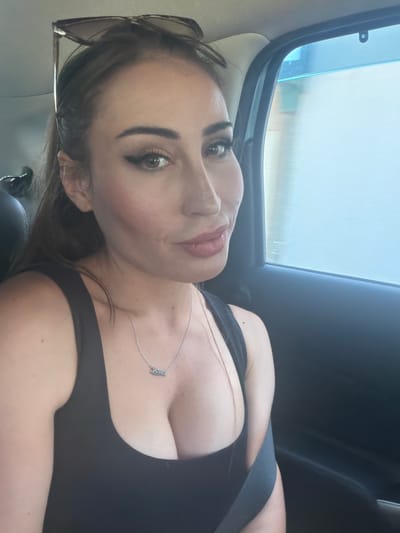 A selfie, taken from a low angle, shows a woman in the backseat of a vehicle. She is wearing a black top and a delicate silver necklace with a nameplate. Sunglasses are perched on her head, and her hair is dark brown, falling around her face. The background shows a car window with a blurred view of the outdoors, suggesting movement. The lighting indicates it is daytime, possibly with overcast skies given the diffused light. The woman's expression is neutral, and the overall mood is casual. Visible text on the necklace reads "Luna". The scene is set in Tserkezoi, Cyprus.