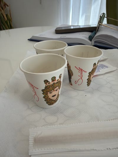 Three disposable white cups, labeled "Sicily" in red script with an image of a woman, sit on a white surface, likely a table, partially covered with paper napkins. A stack of papers or a book with a blue cover is visible behind the cups, along with a pair of glasses. A glimpse of a plant or some other organic material is visible in the lower left corner. The image does not contain any information to confirm the location is in Scordia, Italy.