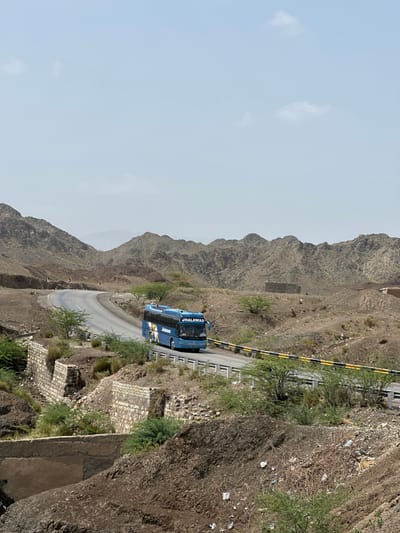 The image shows a blue bus driving along a winding road through a mountainous, arid landscape. The road is paved and has a yellow and black striped barrier along the edge. The mountains are rocky and steep, with sparse vegetation. The sky is overcast, suggesting it might be a cloudy day.
The bus has "JHALAWAN" written on its side in white lettering. The bus appears to be the main object of interest, positioned on the road as it navigates a curve. There are no visible people in the image. The setting is a rugged, natural environment, likely a remote area given the terrain. The overall impression is one of travel through a dramatic landscape. The time of day is unclear due to the overcast sky, but it appears to be daytime. The location cues, such as the mountains and the road, are indicative of a mountainous region. The image captures a moment of transit through this environment.