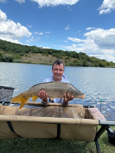 A man in his 40s with glasses and a camouflage-patterned shirt smiles as he holds a large, scale-covered carp horizontally in front of him. The fish's mouth is slightly open, revealing a small barb. The man is positioned in the foreground, in front of a body of water that appears to be a lake or large pond. Behind him, rolling green hills covered in trees stretch across the horizon. The sky above is a vibrant blue, dotted with fluffy white clouds. In the immediate foreground, a tan-colored fishing mat with black straps is visible, and to the left, a black fishing net is partially in view. The scene suggests an outdoor setting, likely a fishing spot in the vicinity of Kolets, Bulgaria.