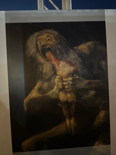 The image shows a painting depicting a monstrous, emaciated figure with long, unkempt hair, hunched over and consuming a severed limb. The figure's mouth is agape, revealing a grotesque tongue and teeth, and its eyes are wide with a manic expression. The scene is dark and atmospheric, with the figures rendered in earthy tones and heavy shadow, suggesting a nightmarish or violent subject matter. The painting appears to be on display, with a portion of a white border and a silver pole visible at the top of the frame, indicating it is likely in a gallery or exhibition. The text at the bottom of the image is partially visible and includes the words "Mazarrón, Spain." This suggests the painting is either located in Mazarrón, Spain, or is an artwork related to the city.