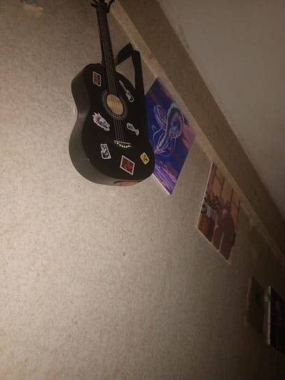 A black acoustic guitar with multiple stickers is mounted on a textured wall. To the right of the guitar, two framed pictures are also hanging on the wall. The picture on the left is a square frame with an abstract design featuring swirling blue and purple lines. The picture on the right is a rectangular frame depicting a vase of flowers casting shadows on a surface. The overall scene appears to be indoors, possibly in a room or hallway, with the lighting suggesting it is either evening or the room is dimly lit. The location is not specified in the image.