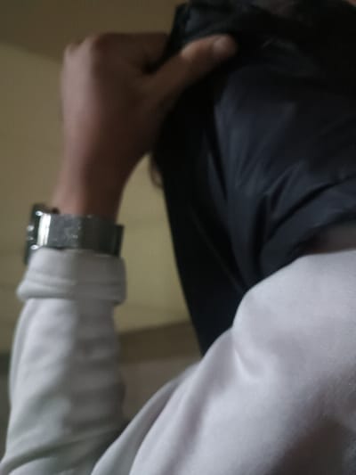 A person's arm and hand are visible. The person wears a wristwatch on their left wrist. The arm is clothed in a light gray or white long-sleeved shirt or sweatshirt. A hand is holding a dark or black piece of cloth. The material of the cloth appears smooth, possibly synthetic, and draped over the person's head. A small portion of skin is visible on the person's finger.