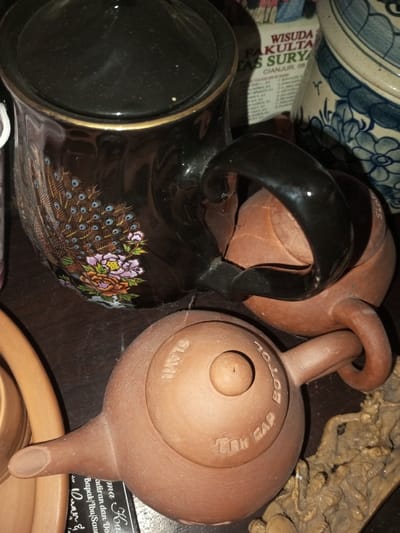 The image shows a collection of ceramic objects. A black ceramic mug with a lid is decorated with a peacock and floral design. Two small, brown teapots are visible, one inscribed with "TEH GAP BOTOL SLAVI." A ceramic jar with blue and white patterns stands in the background, partially obscured. A newspaper clipping and fragments of other items are scattered around the main objects. The scene appears to be a cluttered display on a dark surface.