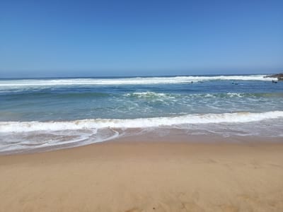 A wide, horizontal, eye-level shot shows a beach in São João das Lampas, Portugal, under a clear, bright blue sky. The foreground is dominated by a sandy beach, with a light brown hue and some faint shadows. The midground features a shoreline where gentle waves are breaking, creating white foam that recedes into the ocean. The water transitions from a clear turquoise near the shore to a deeper blue further out. Several surfers are visible in the water, spread out and appearing to be waiting for waves or paddling. Some are on their surfboards, while others are in the water. In the distance, on the right side of the image, rocky formations protrude from the ocean, and more waves are seen crashing against them. The overall scene conveys a sense of a peaceful day at the beach, likely during the daytime with good weather for surfing. There is no visible text in the image.